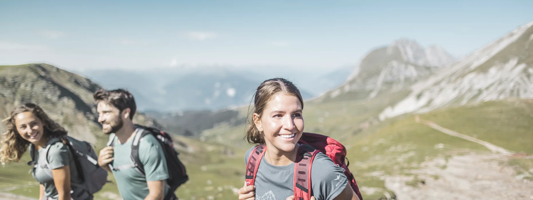 Tre escursionisti sorridenti con zaini su sentiero alpino d'alta quota con tempo soleggiato