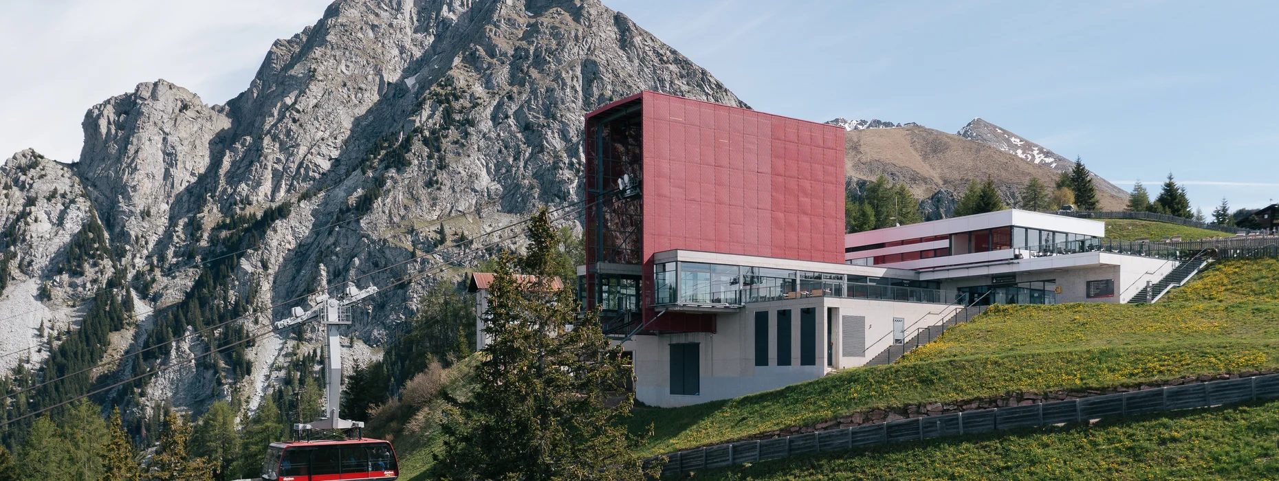 Stazione funiviaria moderna con facciata rossa e cabina davanti a cima rocciosa ripida nelle Alpi