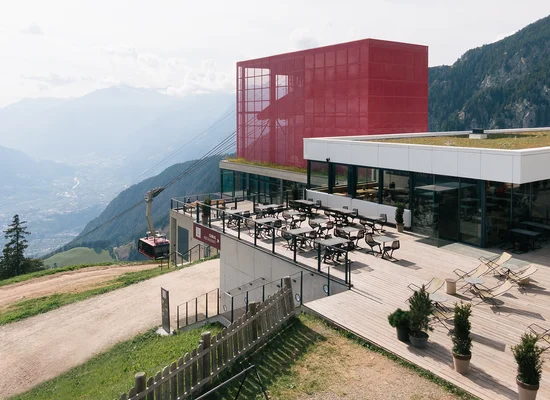 Moderno ristorante di montagna con cubo rosso, terrazza solarium, funivia e panorama alpino in estate