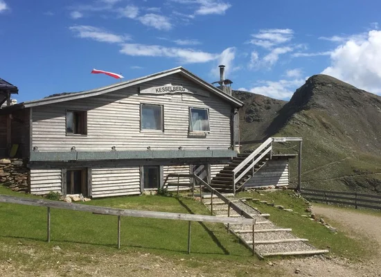 Rifugio Kesselberg in legno con scala esterna, bandiera rosso-bianca e sfondo montano d'estate