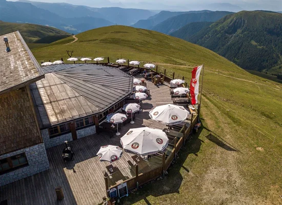 Veduta aerea di malga con grande terrazza in legno, ombrelloni e bandiera sudtirolese