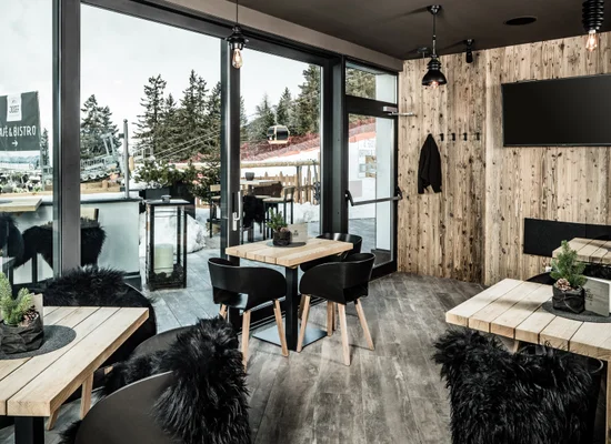 Caffè-bistrot rustico con tavoli in legno, sedie nere, decorazioni in pelliccia e vista sul paesaggio invernale innevato
