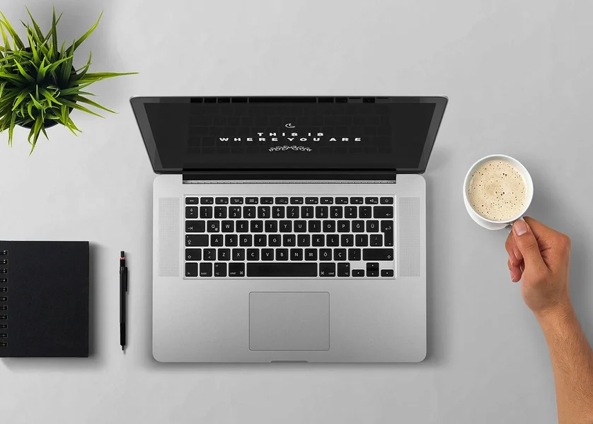 Postazione di lavoro minimalista con laptop, quaderno, pianta e mano che tiene tazza da caffè