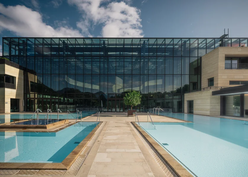 Vista esterna di terme moderne con grande facciata in vetro, due piscine esterne con acqua turchese e un albero al sole