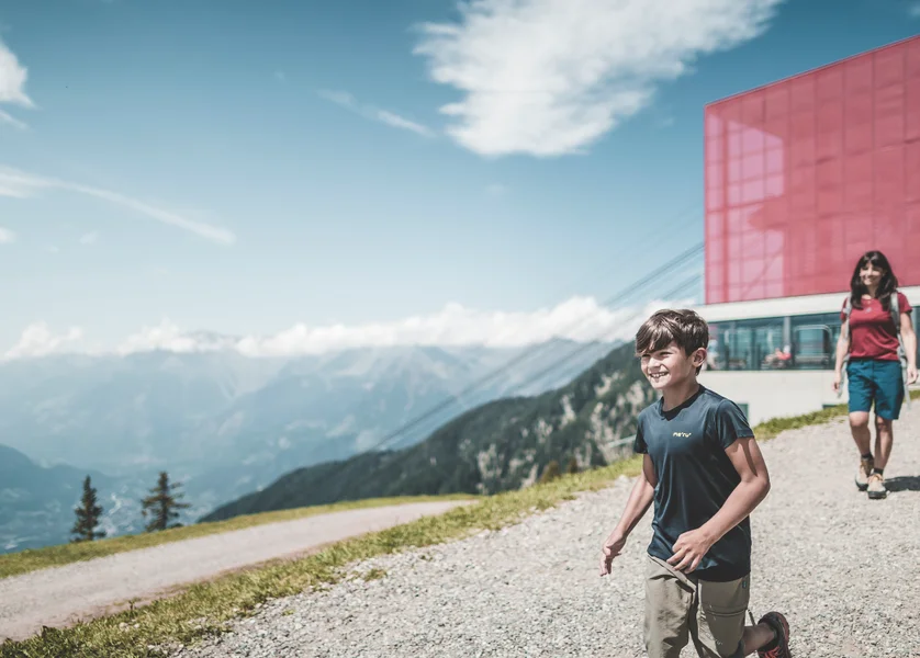 Bambino sorridente con famiglia cammina su sentiero montano vicino a stazione funiviaria con panorama alpino sullo sfondo