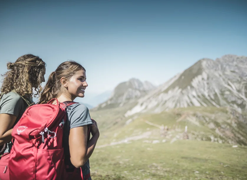Zwei junge Wanderinnen mit Rucksäcken blicken lächelnd auf ein felsiges Alpenpanorama bei klarem Himmel