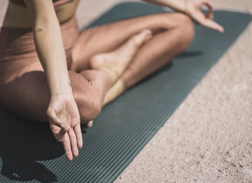 Primo piano di una donna seduta a gambe incrociate su tappetino da yoga verde con gesto mudra e piccoli tatuaggi